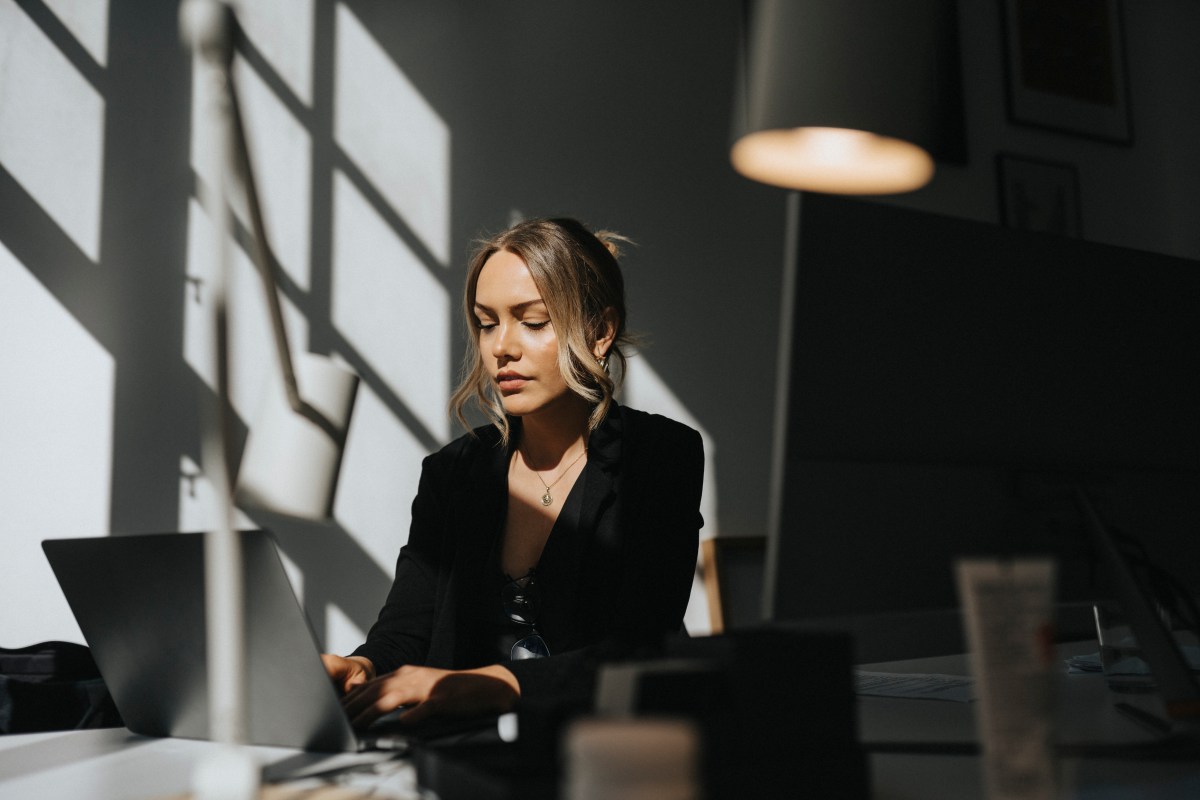 Eine junge Frau mit blonden Haaren sitzt im Büro und arbeitet am Laptop.