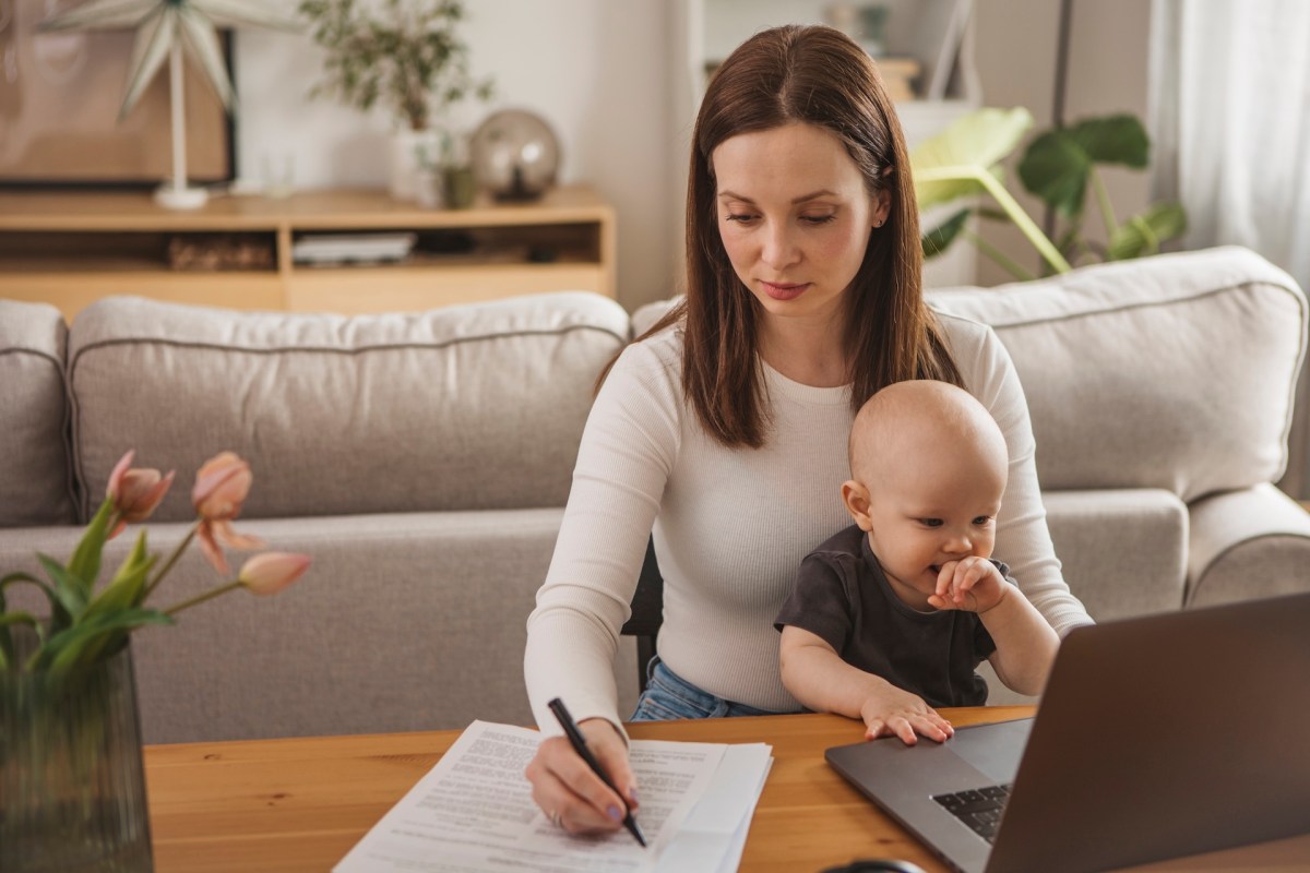 Junge Mutter mit Baby zu Hause, arbeitet am Laptop.