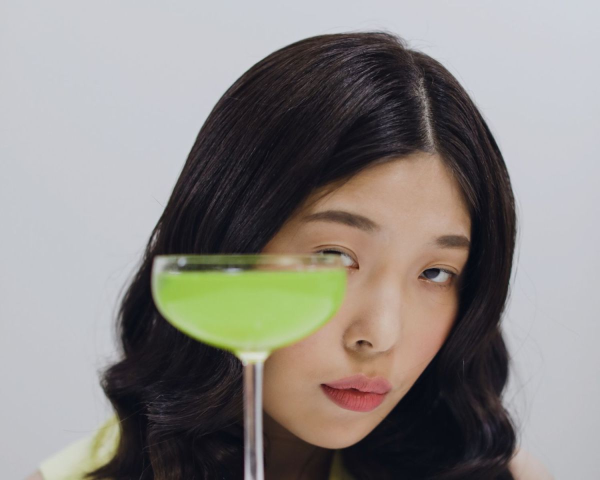 Frau schaut hinter einem mit grüner Flüssigkeit gefülltem Glas hervor.