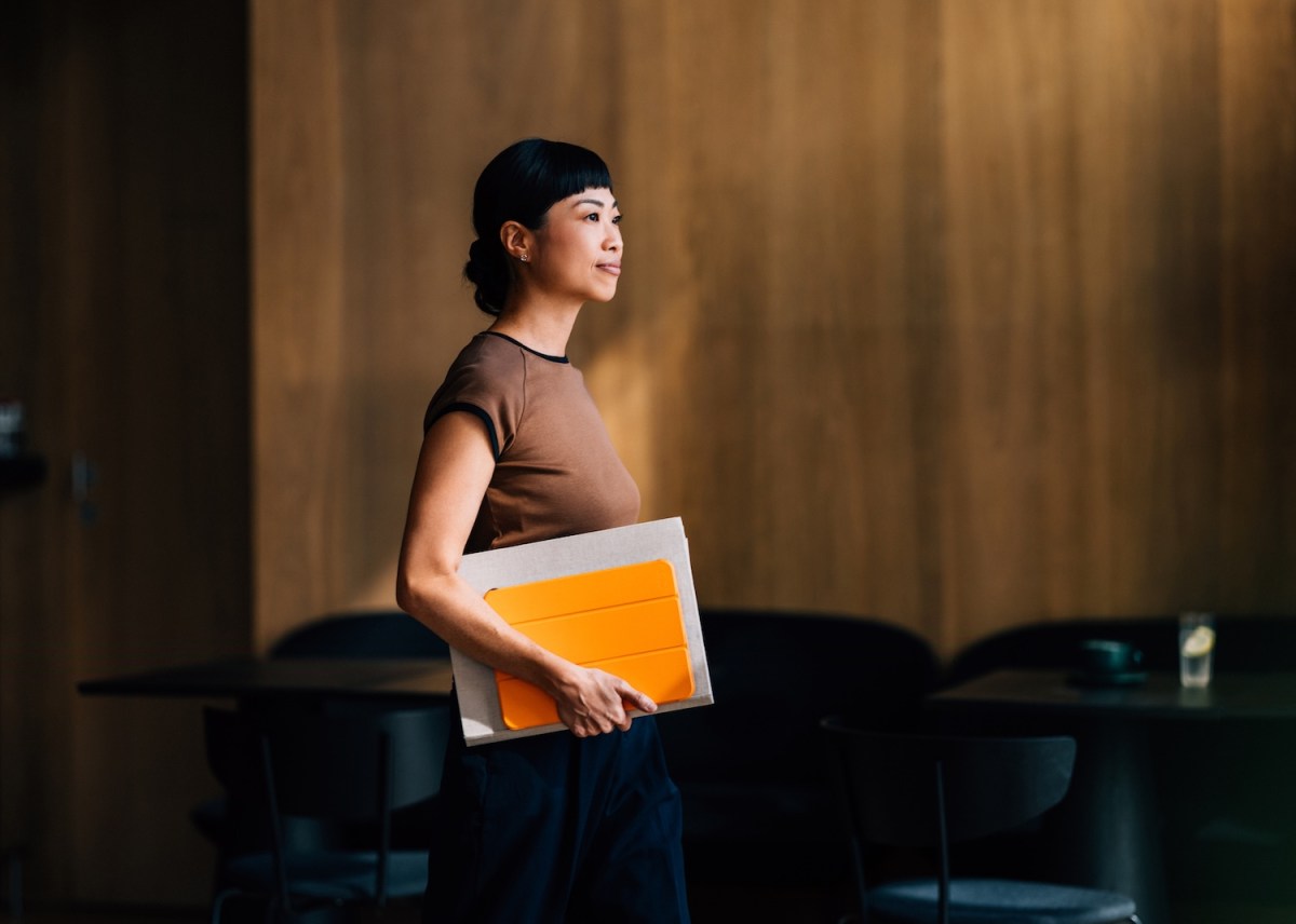 Eine Frau betritt mit einem leuchtend orangefarbenen Notizbuch in der Hand ein modernes Büro.