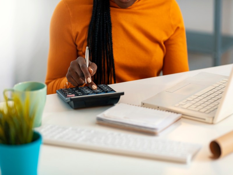 Junge Afroamerikanerin, die mit Laptop und Taschenrechner am Schreibtisch arbeitet.