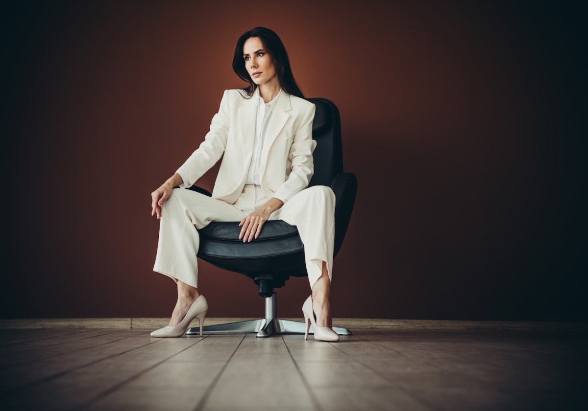 Eine berufstätige Frau im weißen Anzug sitzt selbstbewusst auf einem Stuhl.