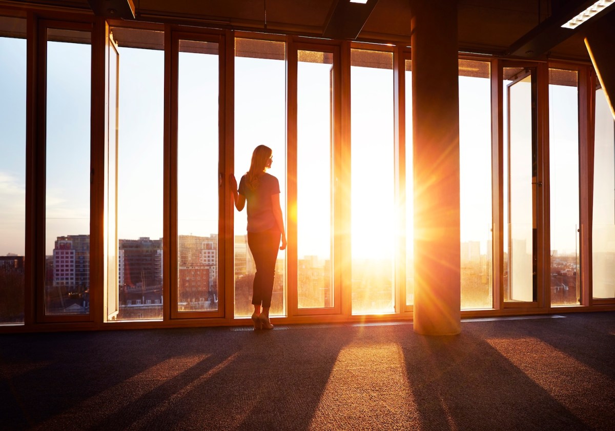 Eine Frau steht im Büro und schaut bei Sonnenuntergang aus dem Fenster.