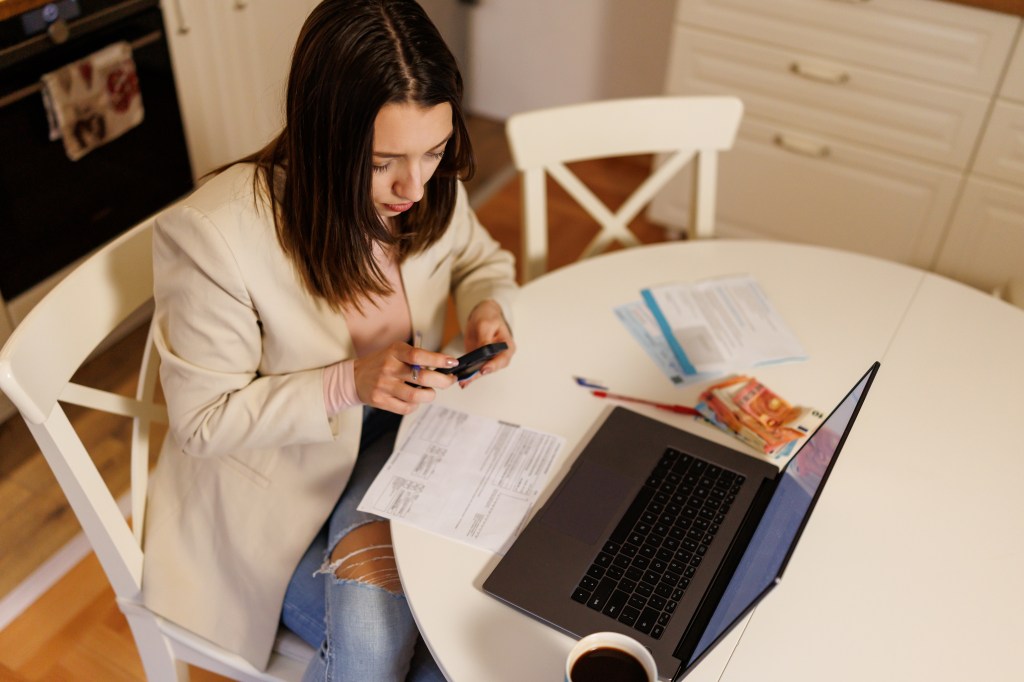 Aufnahme einer jungen Frau aus der Vogelperspektive, die mit ihrem Smartphone und Laptop am Tisch sitzt.