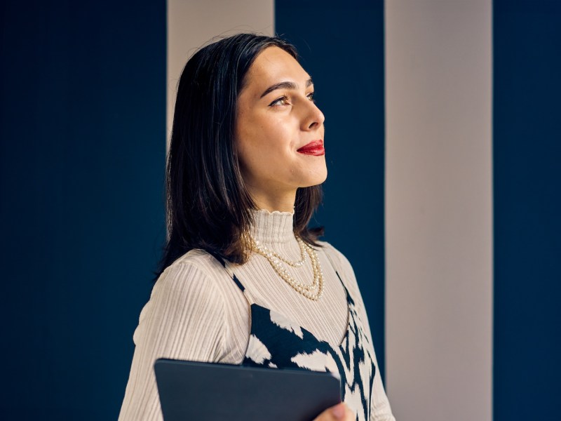 Eine selbstbewusste Frau mit braunen Haaren steht vor einer blau-weiß gestreiften Wand.