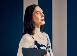 Eine selbstbewusste Frau mit braunen Haaren steht vor einer blau-weiß gestreiften Wand.