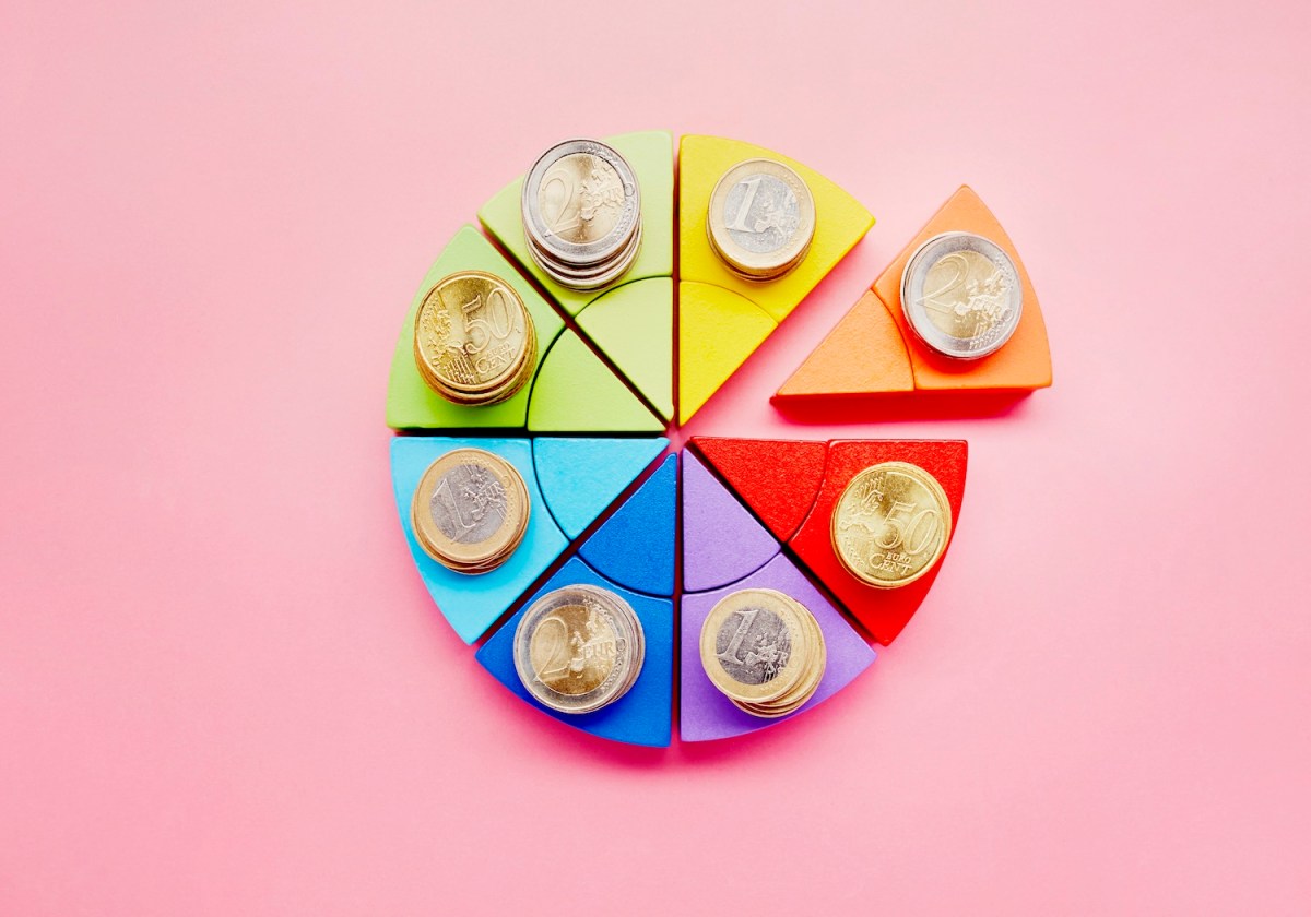 Kreisdiagramm aus bunten Bausteinen und Stapeln von Euro-Münzen auf rosa Hintergrund.