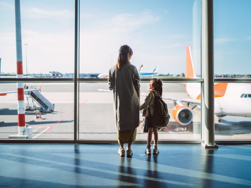 Rückansicht einer Mutter und ihrer kleinen Tochter, die am Fenster eines Flughafenterminals stehen und im Sonnenlicht die Flugzeuge auf dem Rollfeld beobachten.
