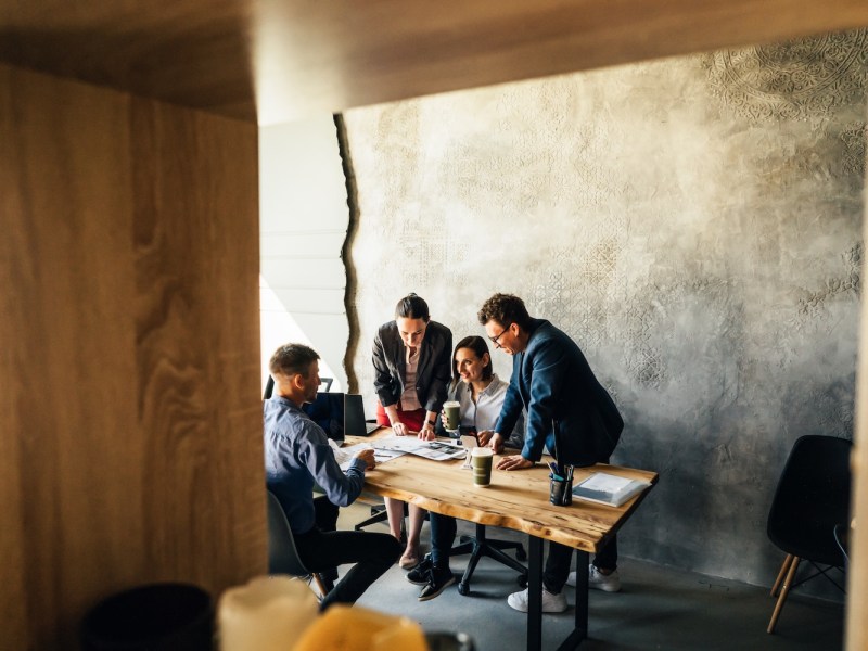 Vier Kollegen sitzen am Tisch im Büro, trinken Kaffee und reden.