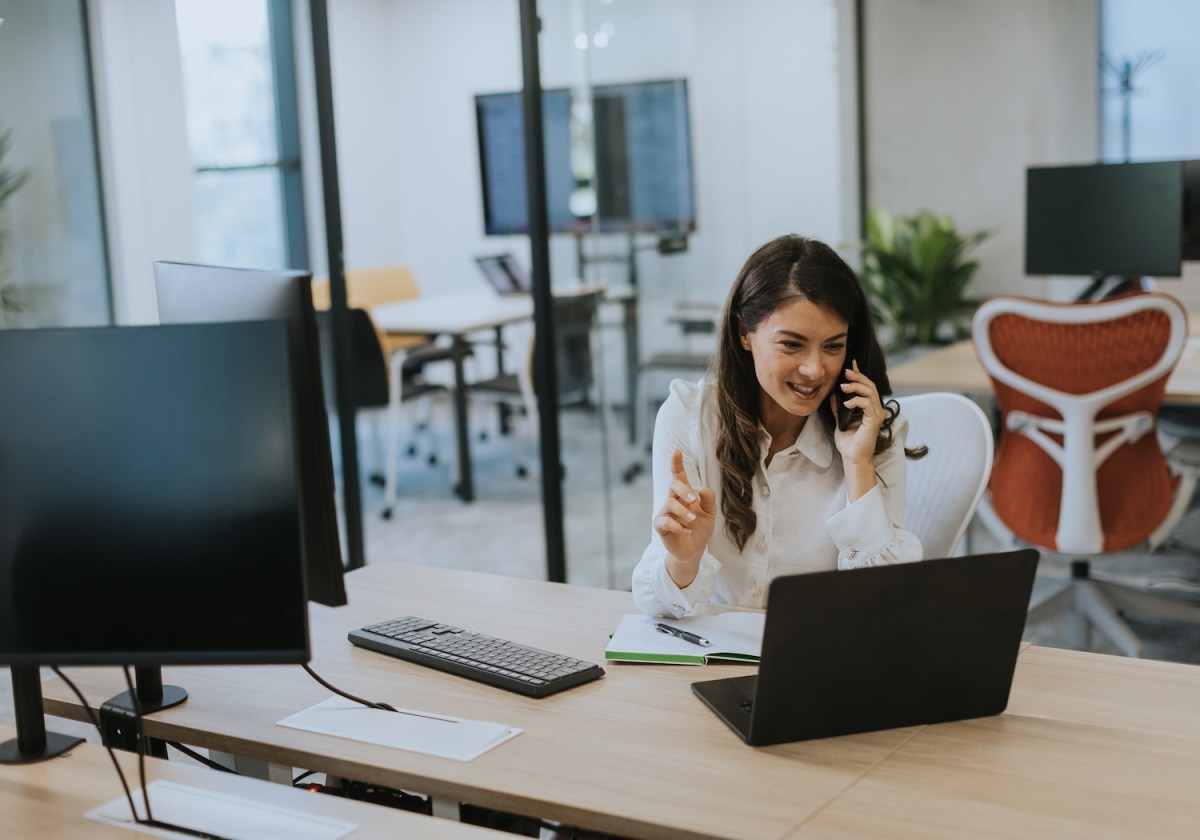Geschäftsfrau, die im modernen Büro mit dem Mobiltelefon arbeitet, während sie am Laptop arbeitet.