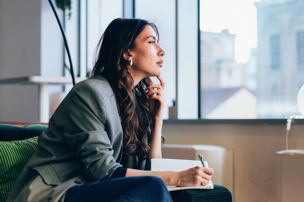 Eine berufstätige Frau sitzt nachdenklich mit Notizbuch und Stift in der Hand in einer modernen Büroumgebung mit großen Fenstern.