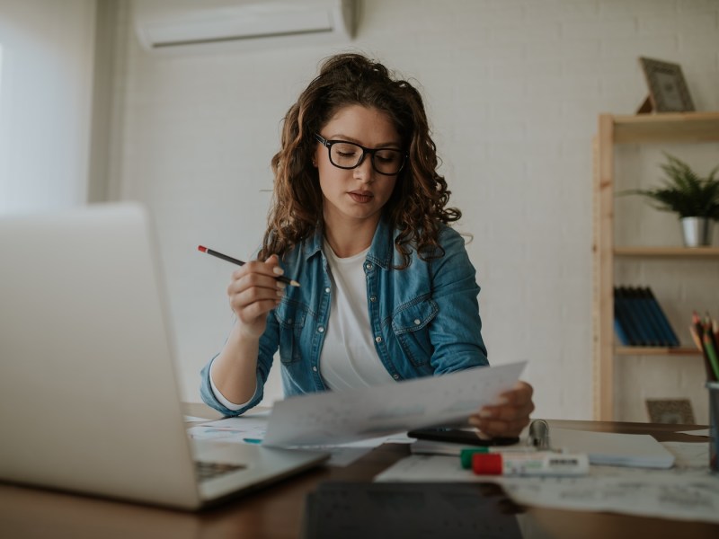 Junge Frau mit Brille im Homeoffice, die sich Papiere ansieht.