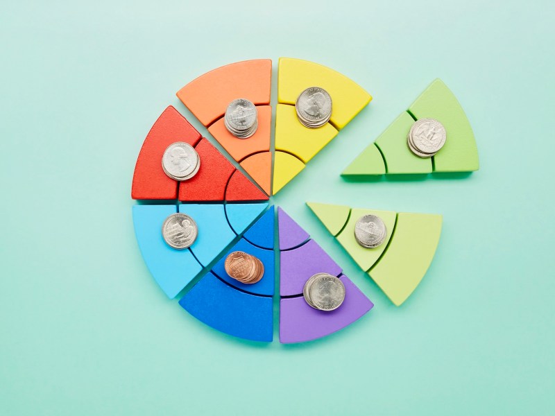 Erhöhte Ansicht eines Kreisdiagramms aus bunten Bausteinen und Stapeln von Münzen auf türkisfarbenem Hintergrund.