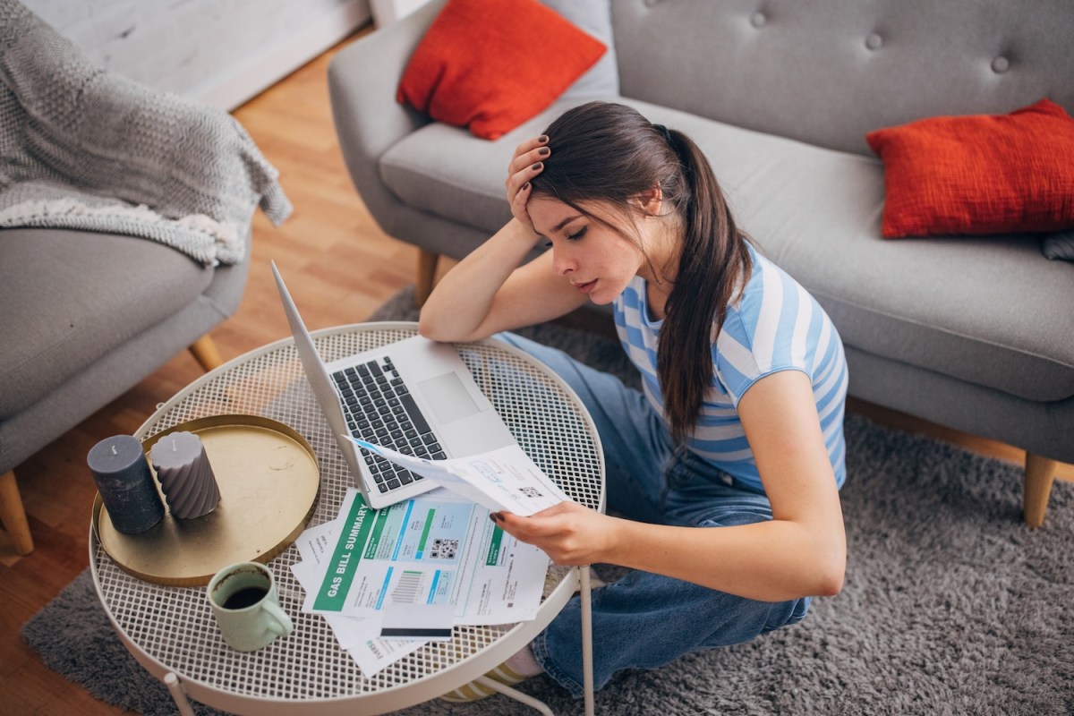 Eine Frau hält nachdenklich den Kopf, während sie in ihrem Wohnzimmer Rechnungen durchgeht.