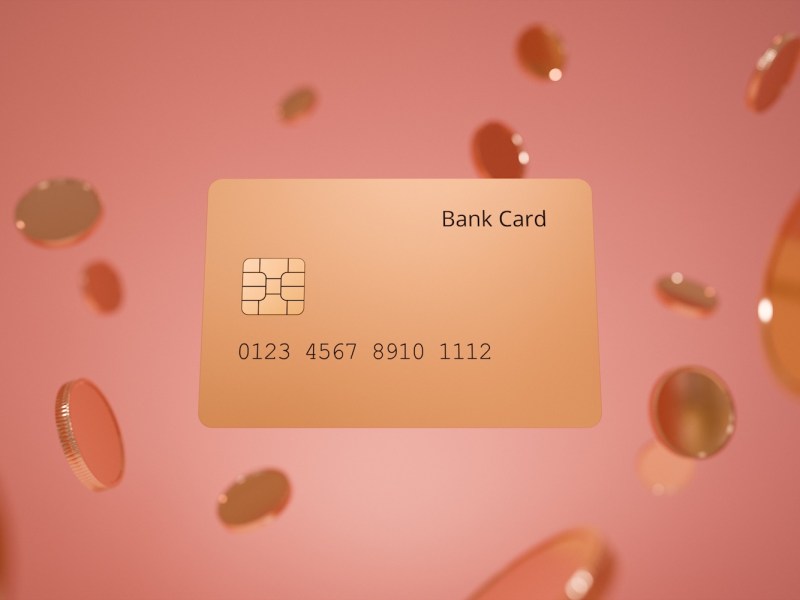 Eine Bankkarte in Goldtönen schwebt zwischen mehreren Münzen vor einem minimalistischen rosa Hintergrund.