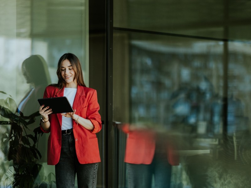 Eine lächelnde, berufstätige Frau steht neben einem modernen Bürogebäude aus Glas, hält ein Tablet in der Hand.