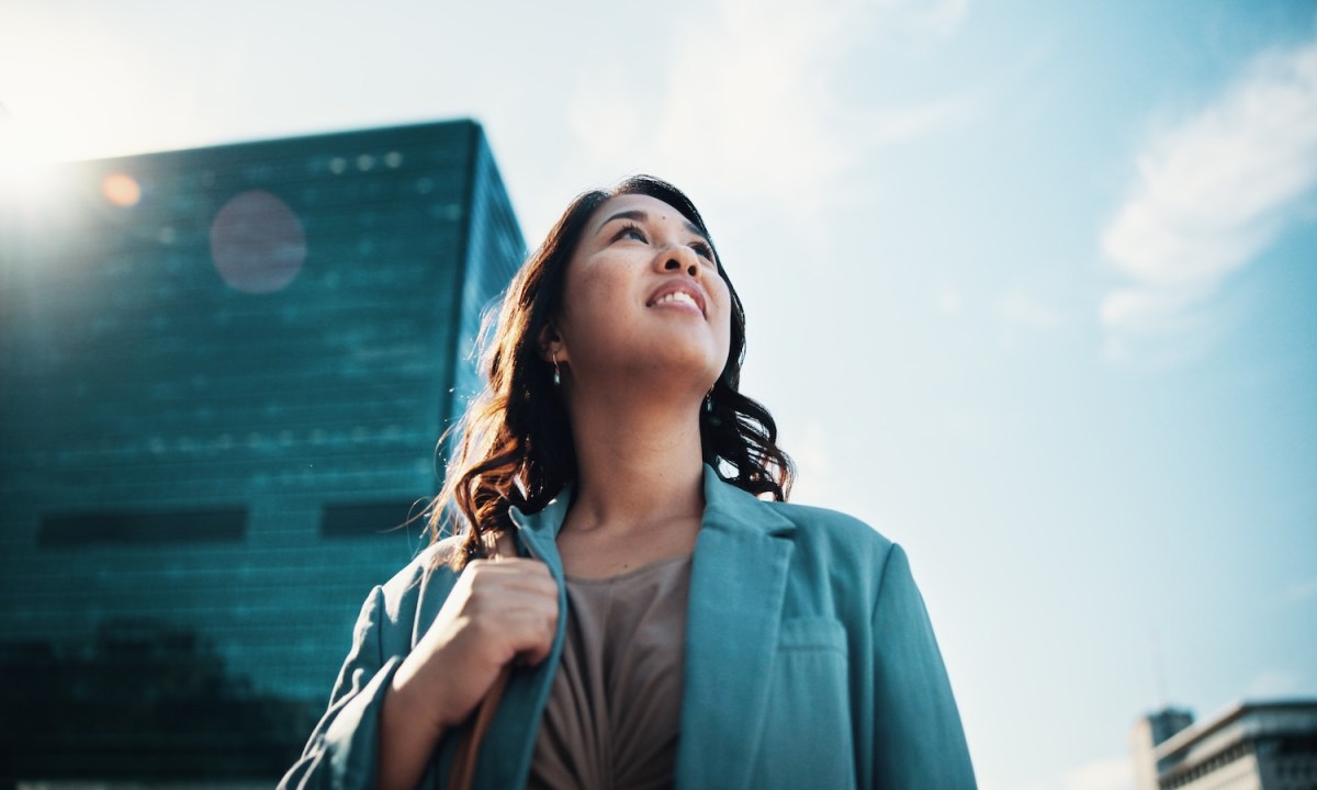 Eine Frau im Anzug schau in den Himmel. Hinter ihr ist ein Bürogebäude zu erkennen.
