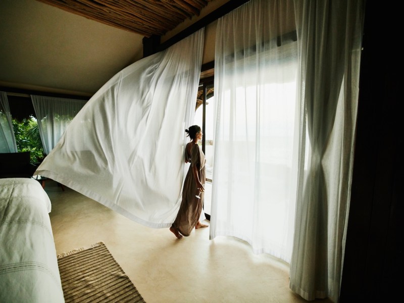 Weitwinkelaufnahme einer Frau, die in einer Luxushotelsuite steht und die Aussicht mit im Wind wehenden Vorhängen genießt.