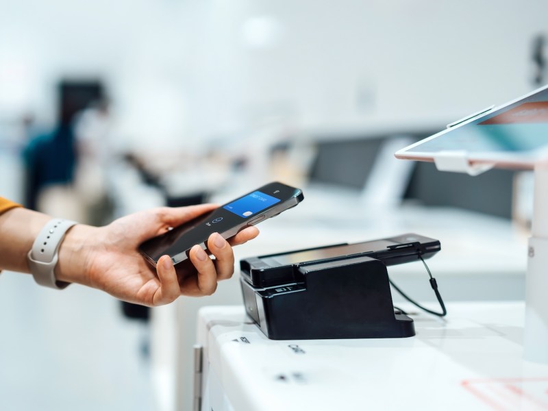 Nahaufnahme einer Frauenhand, die mit ihrem Smartphone kontaktlos an einem Kartenlesegerät in einem Geschäft bezahlt.