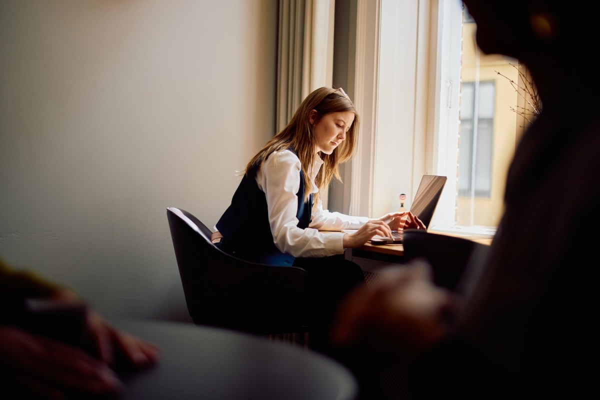 Eine konzentrierte Geschäftsfrau sitzt an einem Fenster und tippt auf ihrem Laptop an einem Holzschreibtisch.
