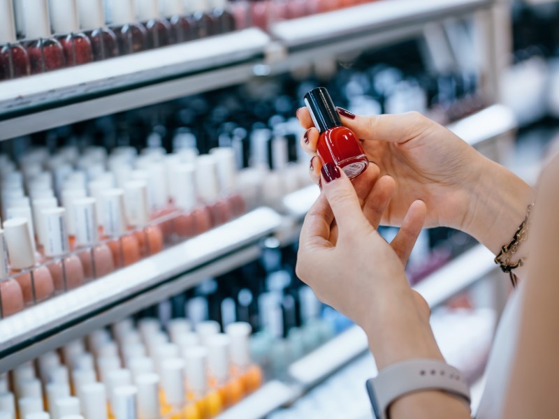 Nahaufnahme einer jungen Frau, die in einem Kosmetikgeschäft einkauft und Nagellack aus einem Regal auswählt.