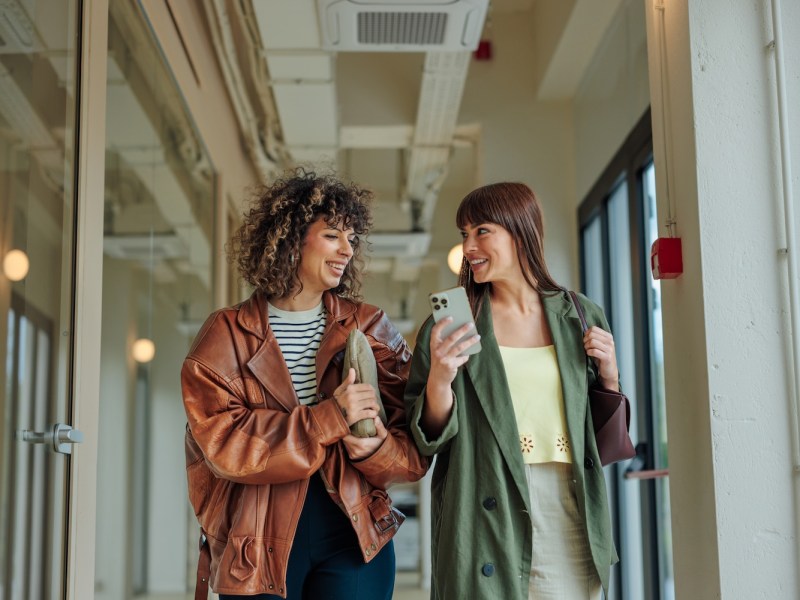 Zwei junge Frauen, vermutlich Kolleginnen, gehen lächelnd und auf ein Smartphone schauend einen Büroflur entlang.