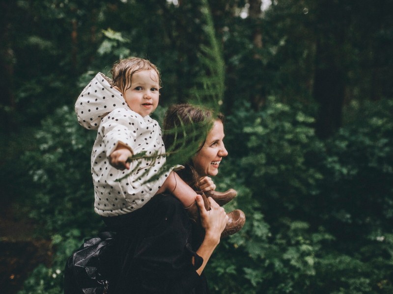 Eine Frau trägt ein Kleinkind auf den Schultern. Sie laufen durch den Wald.