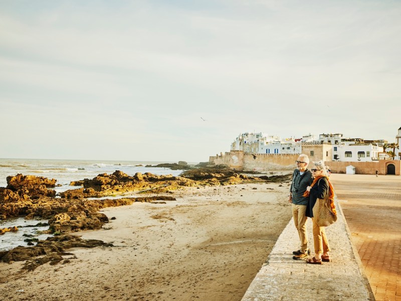 Aufnahme eines älteren Paares, das bei Sonnenaufgang auf einer Ufermauer steht und den Strand und das Meer bewundert.