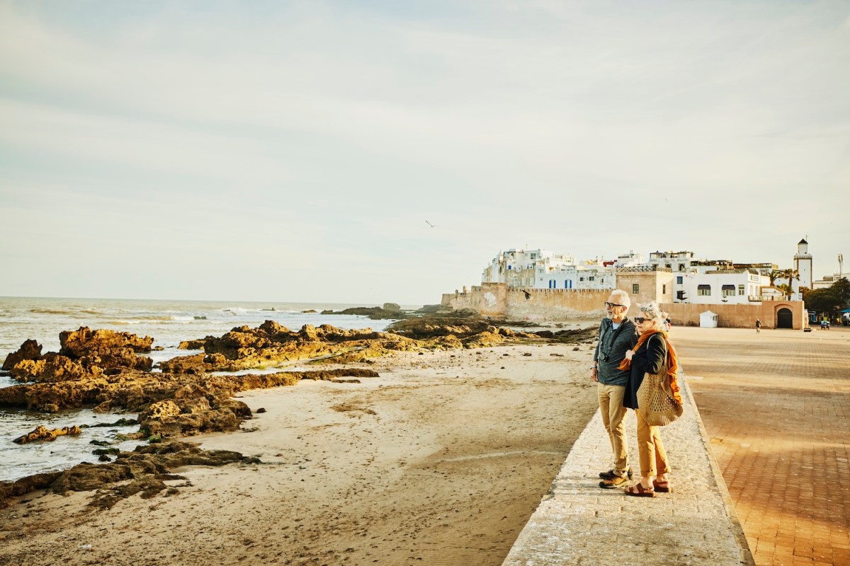 Aufnahme eines älteren Paares, das bei Sonnenaufgang auf einer Ufermauer steht und den Strand und das Meer bewundert.